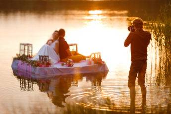 Wedding photographer in action, taking a picture of the bride and groom sitting on the raft. Summer, sunset.