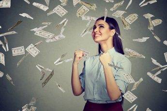 Portrait happy woman exults pumping fists ecstatic celebrates success under a money rain falling down dollar bills banknotes isolated on gray wall background with copy space